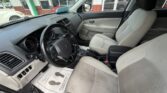 Interior of a light-gray car showing the driver’s seat, steering wheel, center console, and beige front seats at a dealership. Window view includes a storefront outside.