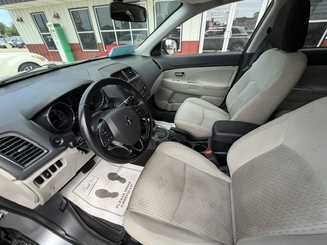 Interior of a light-gray car showing the driver’s seat, steering wheel, center console, and beige front seats at a dealership. Window view includes a storefront outside.