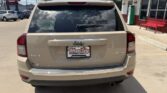 Beige Jeep SUV viewed from the rear in a dealership lot; WheelCity license plate frame, 4x4 badge, single exhaust, storefront to the right.