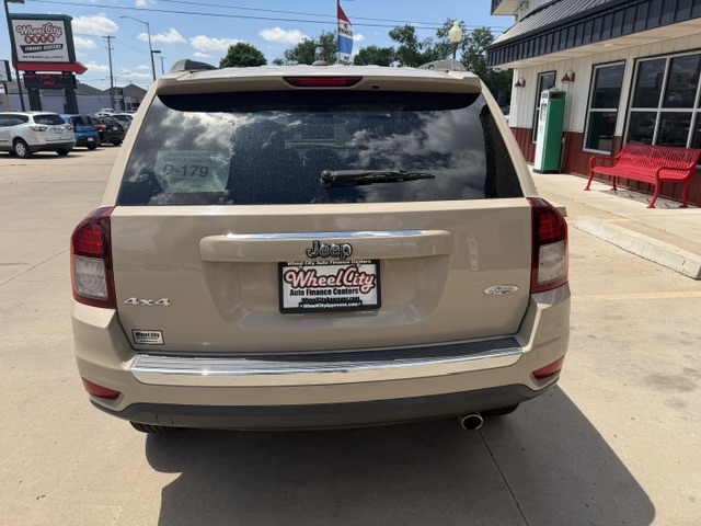 Beige Jeep SUV viewed from the rear in a dealership lot; WheelCity license plate frame, 4x4 badge, single exhaust, storefront to the right.