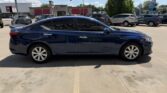 Blue four-door sedan parked in a sunny dealership lot, with other cars and storefronts visible in the background.