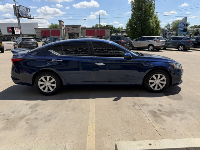 Blue four-door sedan parked in a sunny dealership lot, with other cars and storefronts visible in the background.