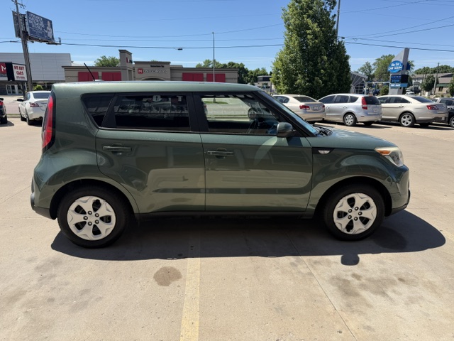 Side view of a green four-door compact hatchback parked in a sunny lot with storefronts in the background.