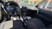 Interior view from the passenger seat of a Nissan car: steering wheel, center console with touchscreen, gear shifter, and cup holders, with dealership lot visible through the windows