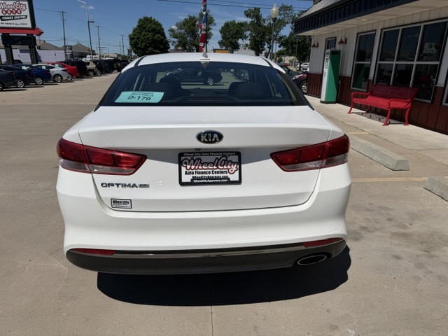 Rear view of a white Kia Optima parked in a dealership lot, with a Wheel City Auto Finance Centers plate frame and a blue sale tag visible on the rear window.