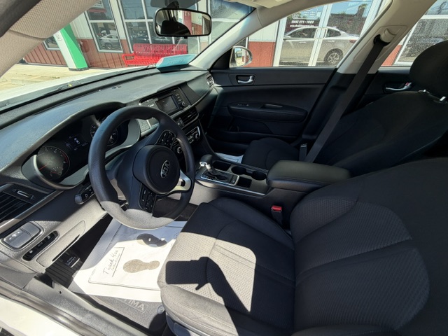 Front interior of a dark cloth sedan: driver’s seat, steering wheel, center console with gear shifter and cupholders, and the front passenger seat; dashboard and windows with sunlight streaming in.