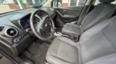 Front interior of a Chevrolet car showing the steering wheel, driver and passenger seats, center console, and dashboard with a blue-lit instrument cluster; view from the open driver's door on a dealership lot behind a building.