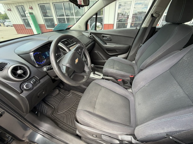 Front interior of a Chevrolet car showing the steering wheel, driver and passenger seats, center console, and dashboard with a blue-lit instrument cluster; view from the open driver's door on a dealership lot behind a building.