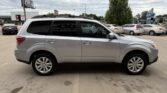 Side view of a silver SUV with roof rails parked in a dealership lot, with other cars and trees in the background.