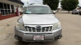 Front view of a silver Subaru SUV parked in a lot outside a storefront, dealership plate visible on the bumper.