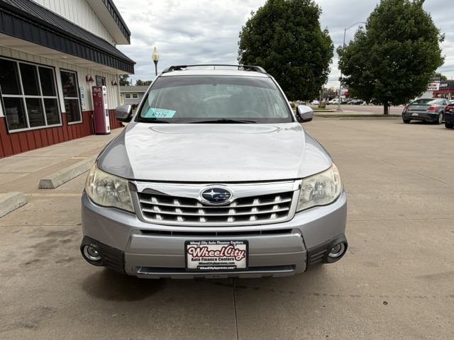 Front view of a silver Subaru SUV parked in a lot outside a storefront, dealership plate visible on the bumper.