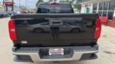 Rear view of a black pickup truck in a car dealership lot; chrome bumper, red taillights, and a Wheel City license plate frame visible on the tailgate.