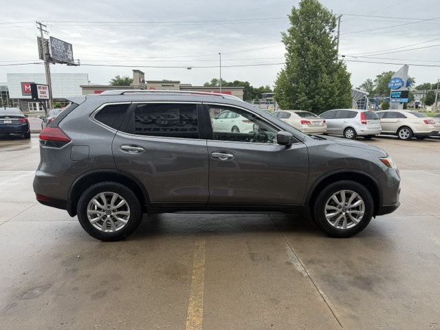 Side view of a gray mid-size SUV parked in a car dealership lot, with other vehicles and storefronts in the background during daytime.