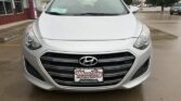 Front view of a silver Hyundai at a dealership, with a Wheel City Auto license plate, on wet pavement with storefronts and trees in the background.