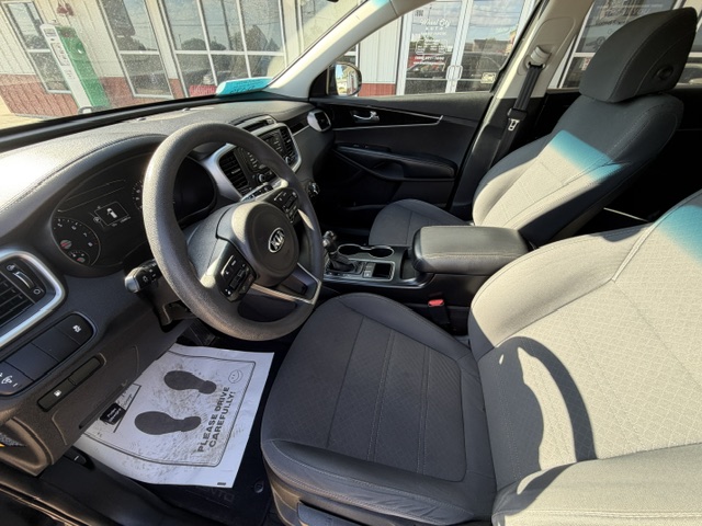 Interior of a modern SUV: steering wheel, dashboard, center console, and gray cloth front seats visible from the driver’s side window. A floor mat with footprints is on the driver's side. Outside scenery seen through the windshield.