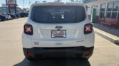 Rear view of a white Jeep SUV parked in a dealership lot, displaying square taillights, black bumper, and a Wheel City license plate frame near a storefront.