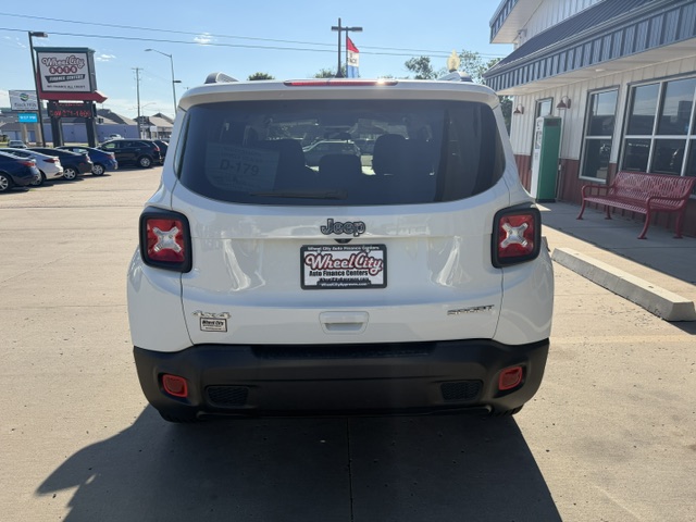 Rear view of a white Jeep SUV parked in a dealership lot, displaying square taillights, black bumper, and a Wheel City license plate frame near a storefront.