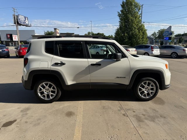 Side profile of a white Jeep Renegade SUV parked in a sunny parking lot, with other cars and a few buildings in the background.