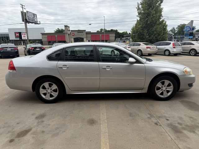 Side profile of a silver four-door sedan parked in a shopping plaza parking lot with storefronts in the background.