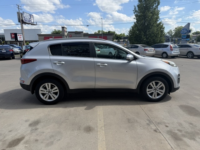 Side view of a silver 4-door SUV in a dealership lot, with neighboring cars and a blue sky above.