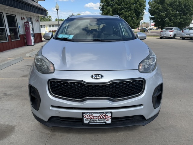 Front view of a silver Kia SUV parked in a car dealership lot, with a Wheel City license plate frame visible on the bumper and a red building to the left.