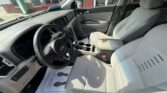Interior of a light-gray Kia vehicle: driver and front passenger seats, dashboard, steering wheel, center console with cup holders, and sunlit front cabin visible through the open door.