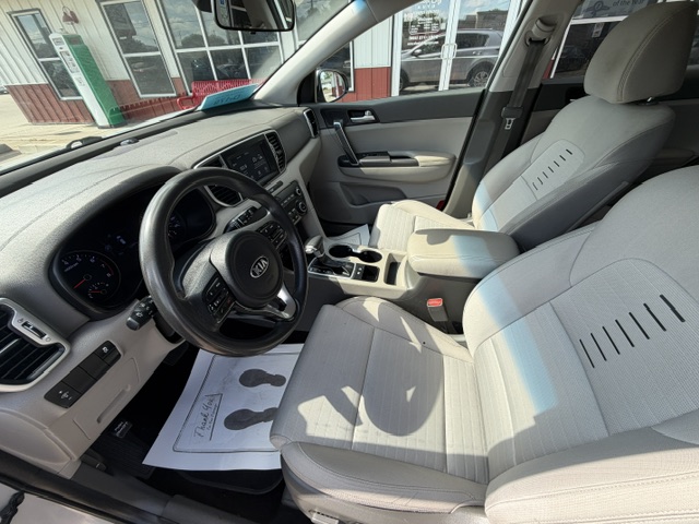 Interior of a light-gray Kia vehicle: driver and front passenger seats, dashboard, steering wheel, center console with cup holders, and sunlit front cabin visible through the open door.