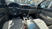 Interior of a Kia sedan showing the steering wheel, center touchscreen, climate controls, gear shifter, cup holders, and light gray fabric seats; sunny day visible through the windows.