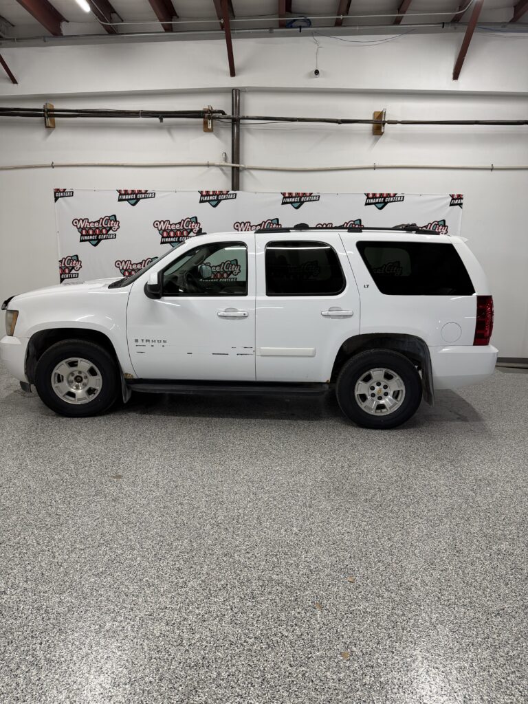Side view of a white Chevrolet Tahoe LT parked indoors against a Wheel City banner backdrop in a showroom