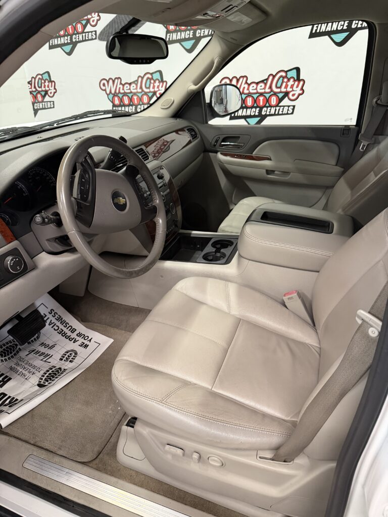Beige leather interior of a Chevrolet SUV, showing the steering wheel, dashboard, center console, and front passenger seat with a dealer backdrop outside the window.