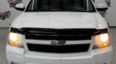 Front view of a white Chevrolet SUV with headlights on, showroom backdrop featuring Wheel City Auto Finance Centers logos.