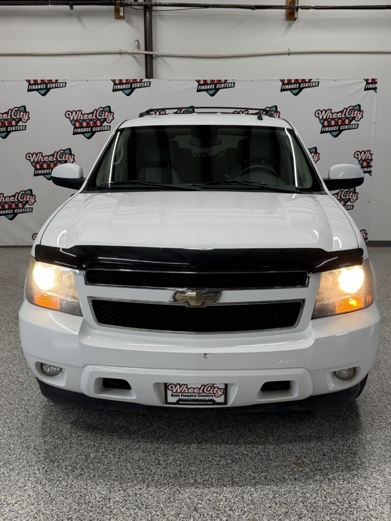 Front view of a white Chevrolet SUV with headlights on, showroom backdrop featuring Wheel City Auto Finance Centers logos.