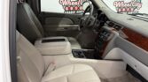 Interior view of a beige-leather car cabin with driver’s seat, steering wheel, center console, and front passenger seat visible. Outside the window are dealership signs reading Wheel City Auto.
