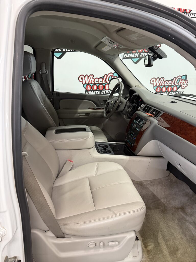 Interior view of a beige-leather car cabin with driver’s seat, steering wheel, center console, and front passenger seat visible. Outside the window are dealership signs reading Wheel City Auto.