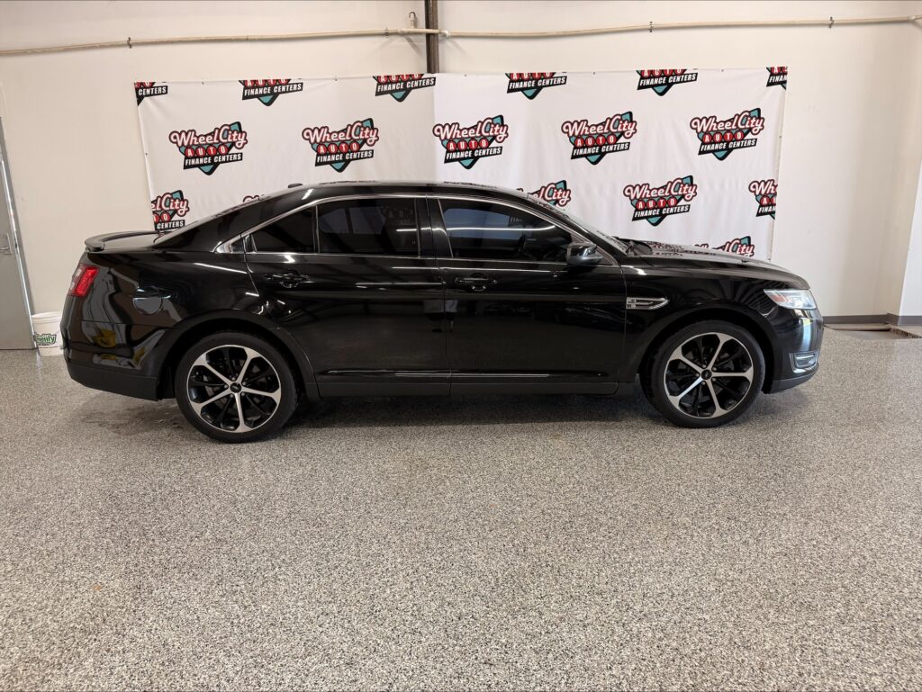 Black sedan parked indoors in front of a Wheel City Auto banner wall showroom backdrop, side profile view.