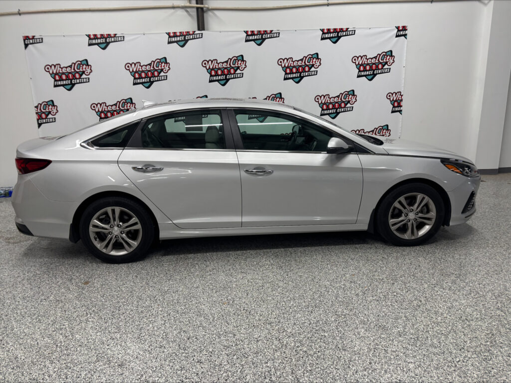 Side profile of a silver sedan parked indoors in front of a Wheel City Auto Finance Centers backdrop banner.