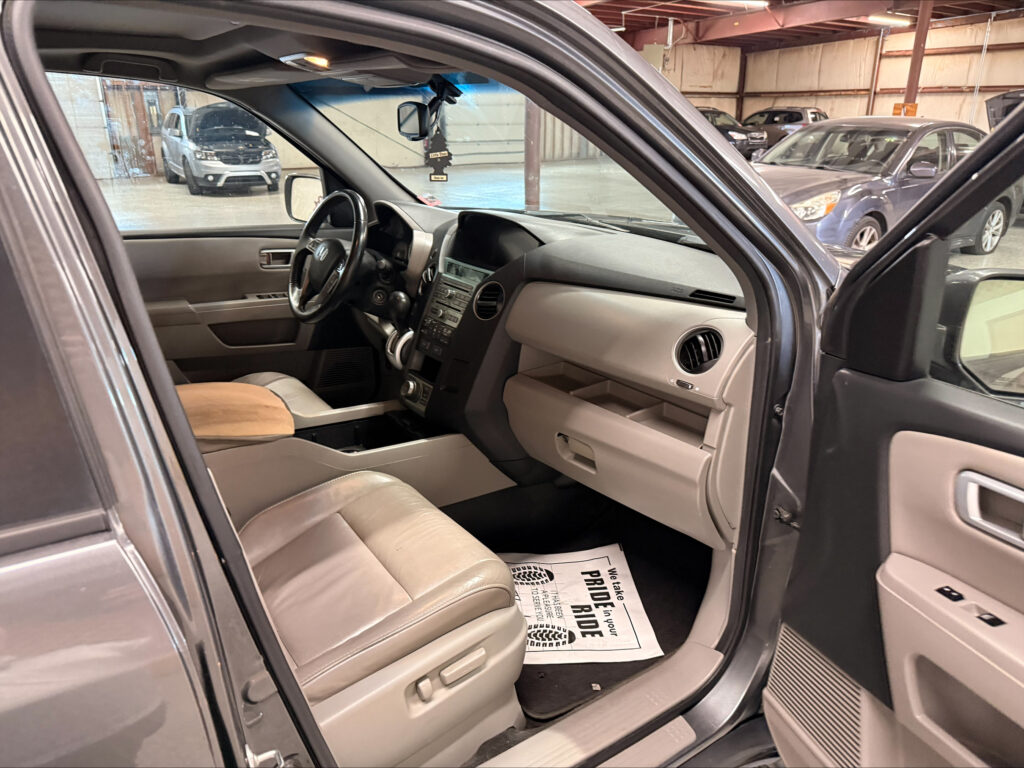 Open front passenger door of a vehicle showing beige leather seats and dashboard, parked in a garage with other cars in the background.
