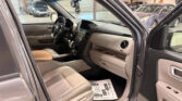Open front passenger door of a vehicle showing beige leather seats and dashboard, parked in a garage with other cars in the background.