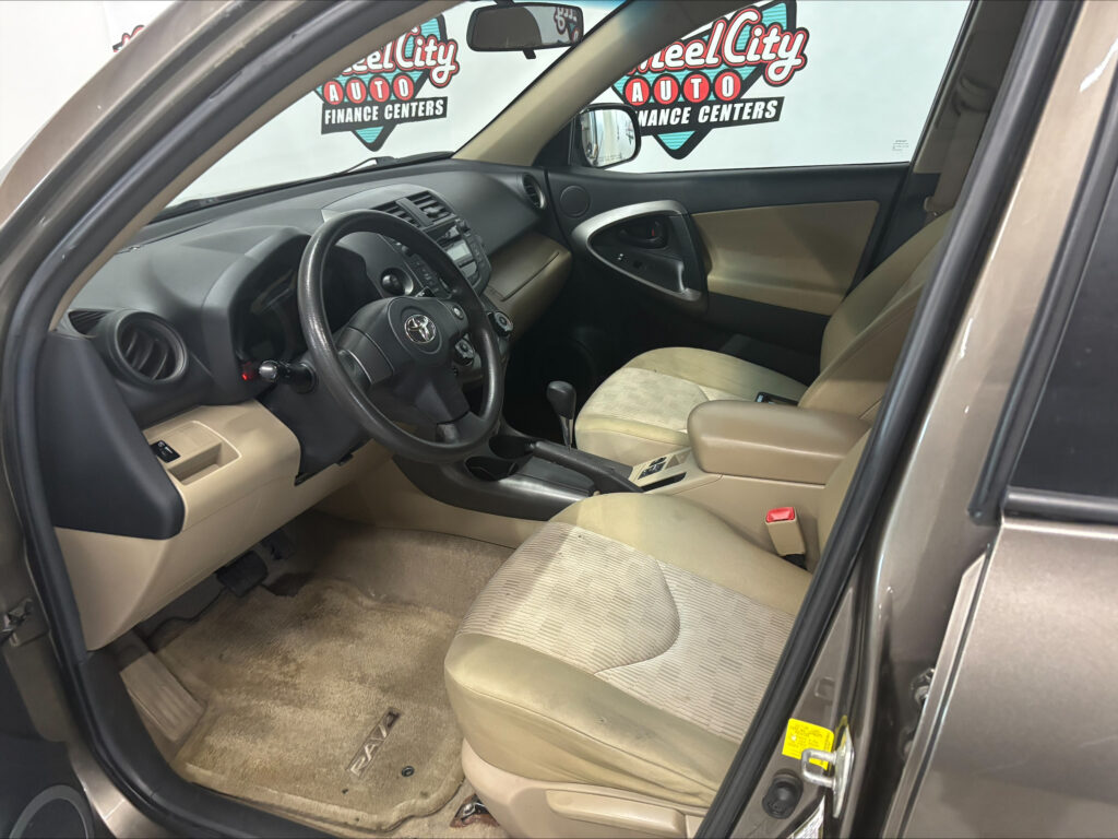 Interior of a beige Toyota SUV: steering wheel, dashboard, center console, driver and front passenger seats, with 'RAV4' floor mat visible