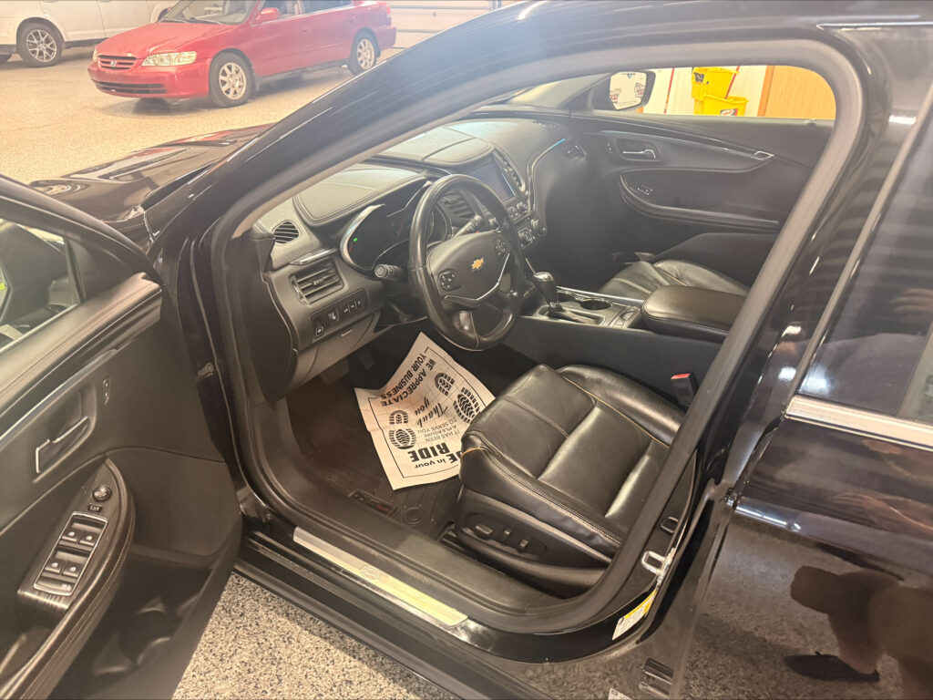 Interior of a black SUV with the driver's door open, showing leather front seats, steering wheel, and center console; paper mat on the floor; showroom with a red car in the background.