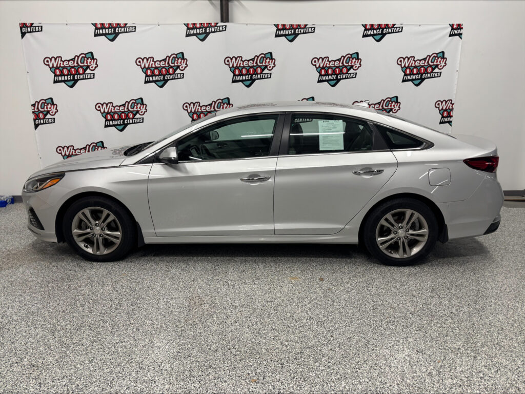 Side view of a silver sedan parked indoors in front of a Wheel City Auto Finance Centers backdrop banner(s)