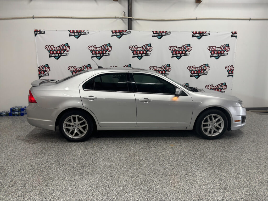 Side profile of a silver sedan parked indoors with a Wheel City Auto Finance Centers banner backdrop behind it.