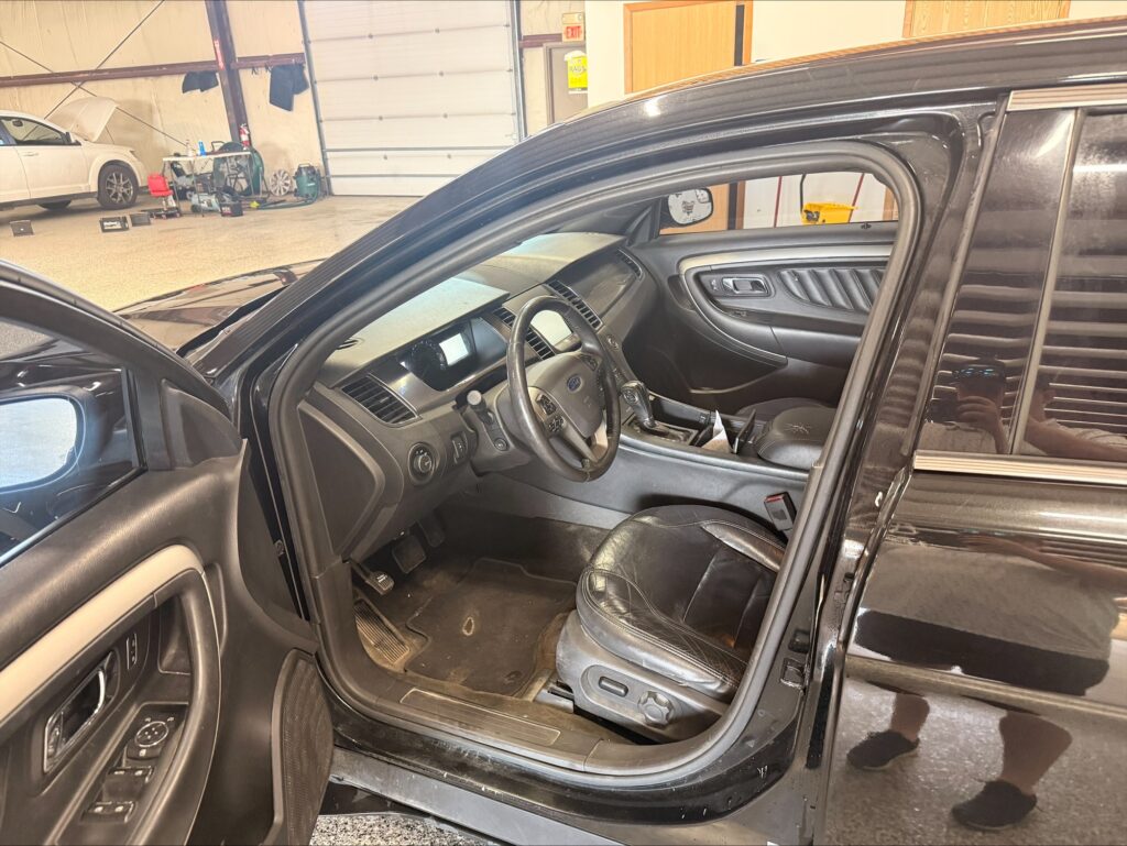 Open front driver’s door of a black SUV, showing the steering wheel, dashboard, and leather seat with a workshop and a white car in the background.