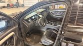 Open front driver’s door of a black SUV, showing the steering wheel, dashboard, and leather seat with a workshop and a white car in the background.