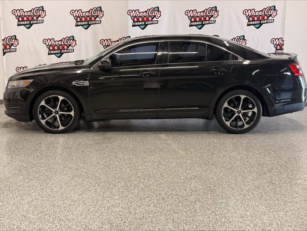 Black four-door sedan in a showroom with a Wheel City Auto Finance Centers backdrop behind it, side view showing alloy wheels and tinted windows.
