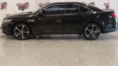 Black four-door sedan in a showroom with a Wheel City Auto Finance Centers backdrop behind it, side view showing alloy wheels and tinted windows.