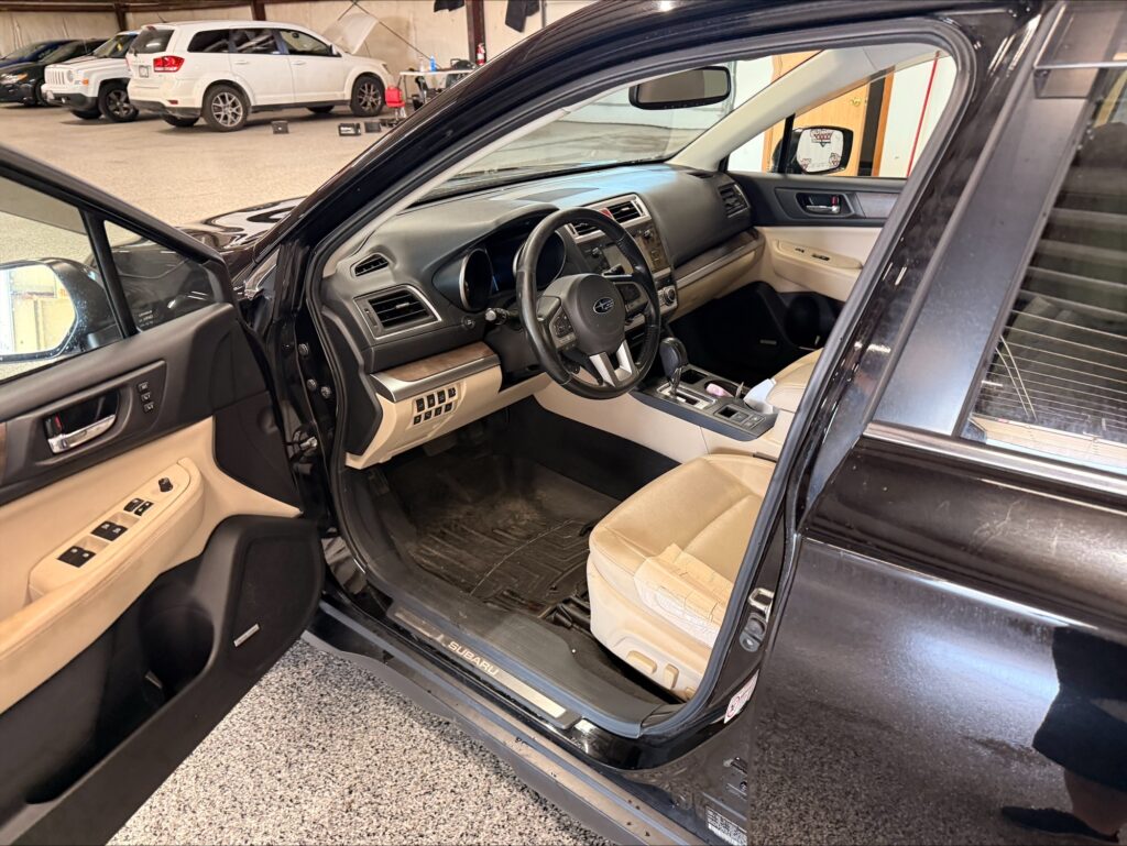 Inside a beige Subaru SUV with the driver’s door open, showing the steering wheel, dashboard, and front seats in a showroom.