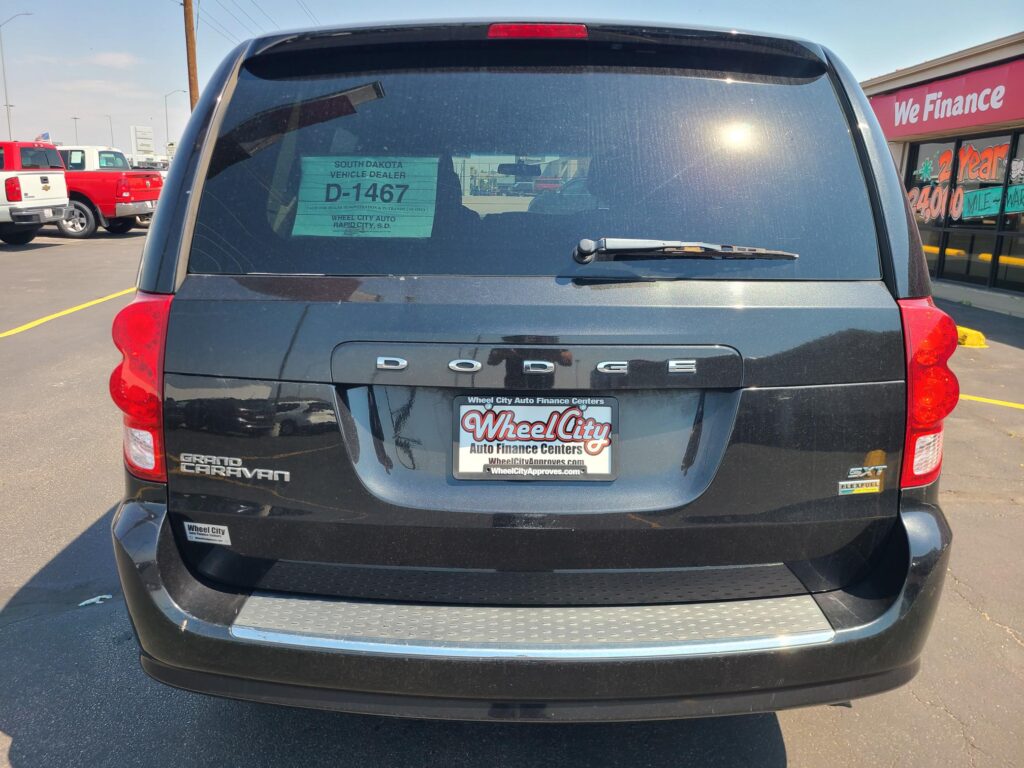 Rear view of a black Dodge Grand Caravan in a dealership lot, with a green D-1467 window sticker and Wheel City license plate frame.