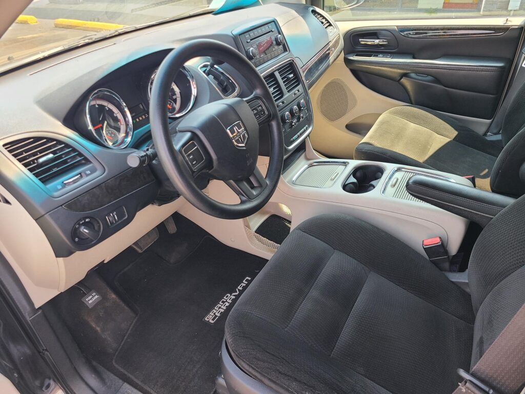 Front cabin of a Dodge Grand Caravan: steering wheel, beige dashboard, center console, and front seats with a floor mat reading 'Grand Caravan'.