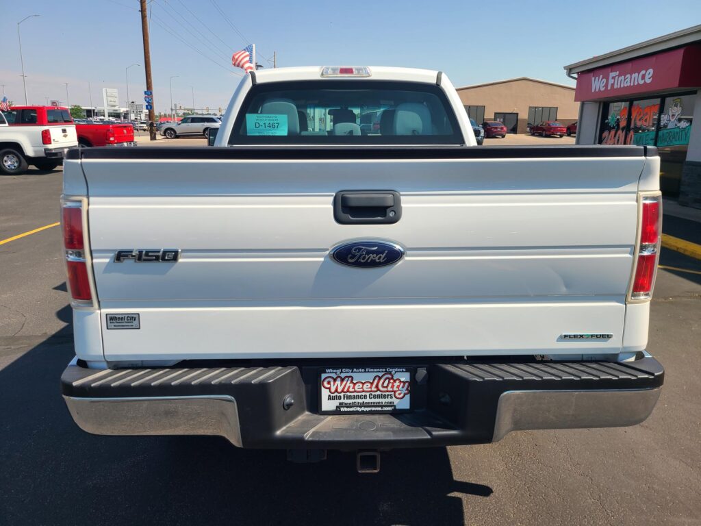 Rear view of a white Ford F-150 pickup in a dealership parking lot, with a Wheel City license plate frame visible in the bumper area.